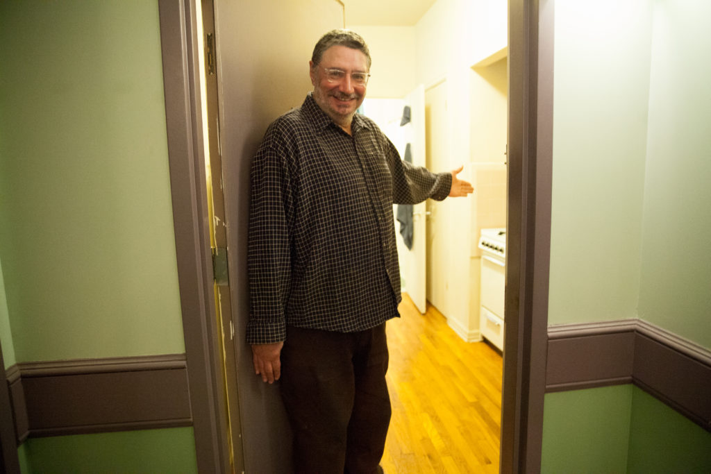 A man standing in front of a apartment door with his hand extended out to the door to invite you inside the room.