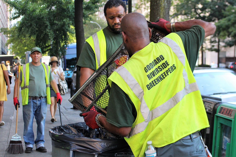 Two Goddard Riverside Green Keepers in bright green vests empty a garbage receptacle on an Upper West Side street.
