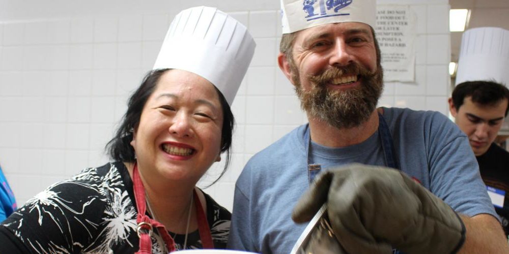 Two longtime volunteers wearing white paper chef's toques smile while preparing food for the  Holiday Meals 