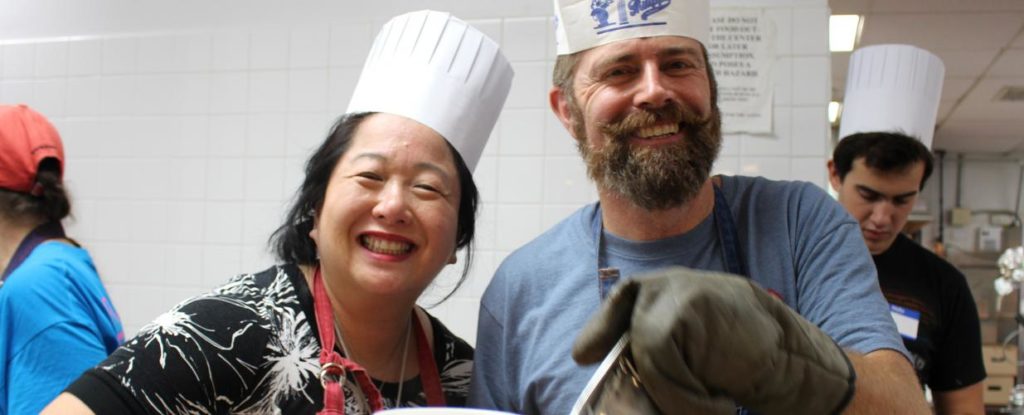 A woman and a man wearing paper chef's toques smile at the camera while cooking food for the Holiday Meals