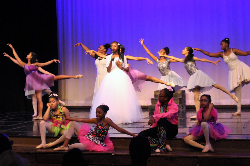 A group of young girls dancing ballet in the background with a young girl singing and another group of girls sitting on the edge of the stage during the Performing Arts Conservatory's final performance in 2018.