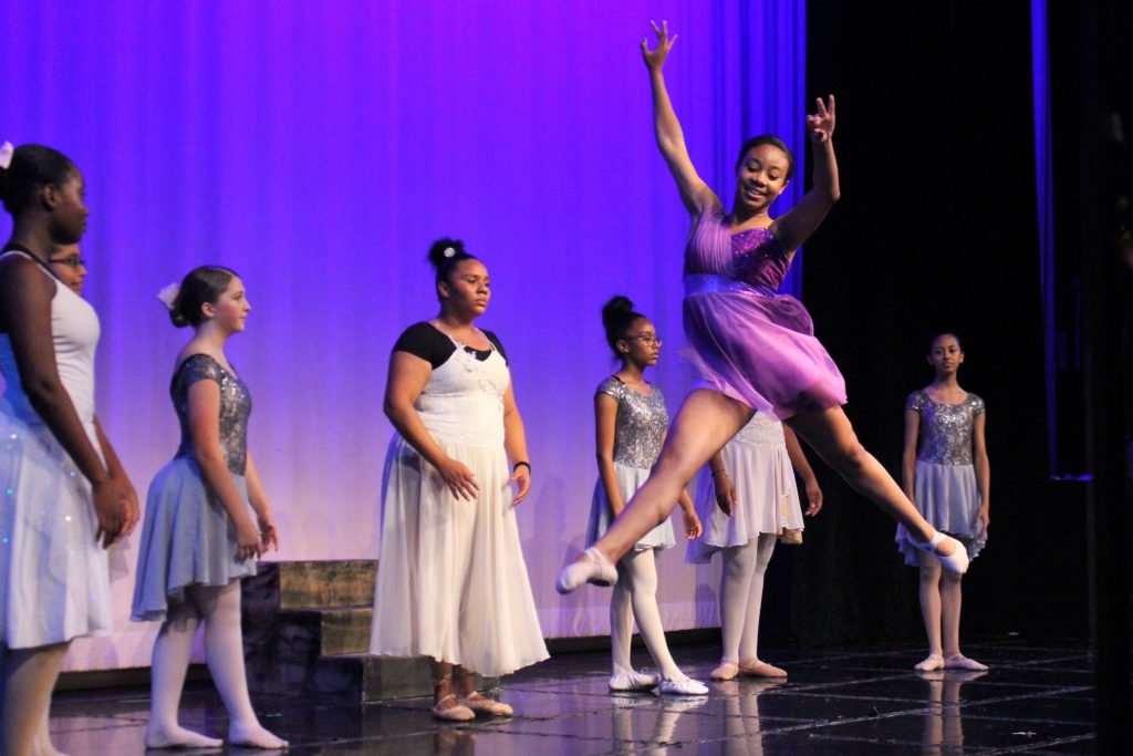 A young girl dancing ballet in front of a group of girls standing behind her on the stage during the Performing Arts Conservatory's final performance in 2018.