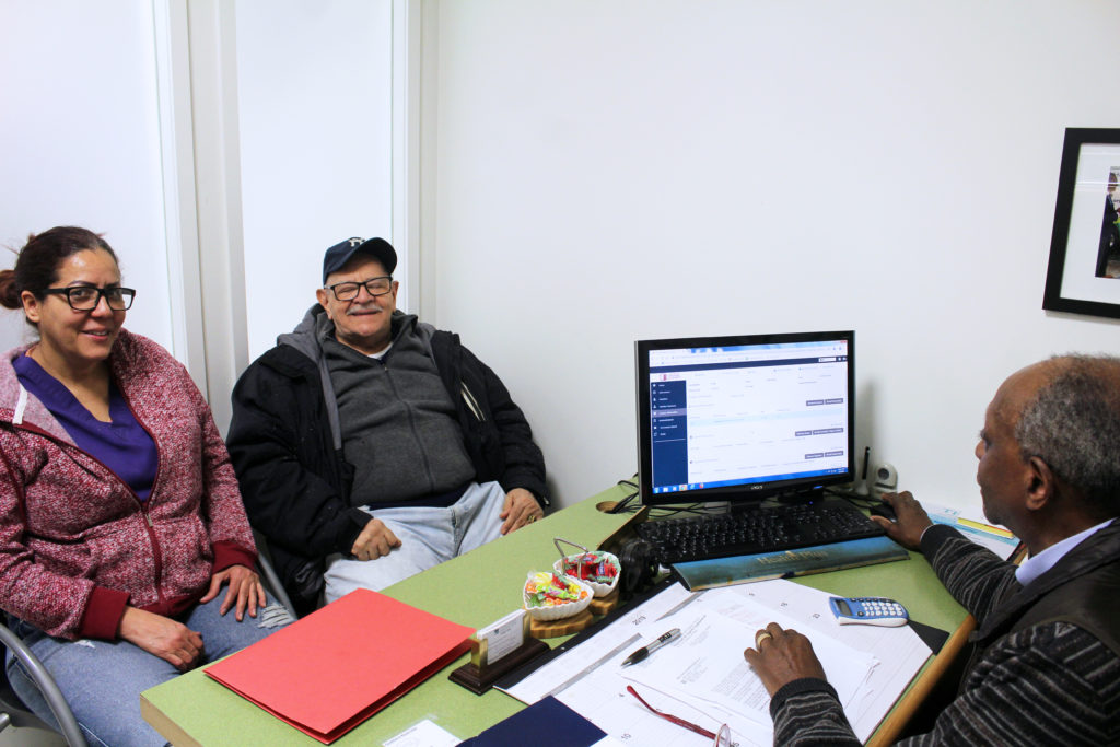 A man and woman sitting in front of an office desk with a man in front of his computer while talking to them at the Resource Center.