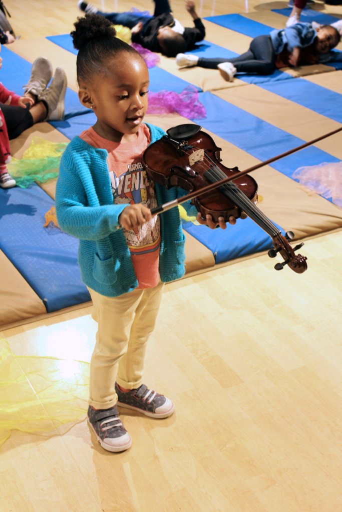A little girl holding a small violin with a floor mat with children laying down in the background.