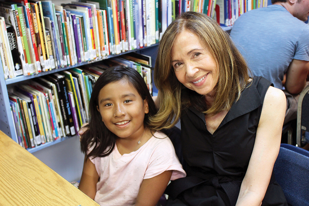 A woman and young girl smiling while seated at a desk next to a bookshelf.