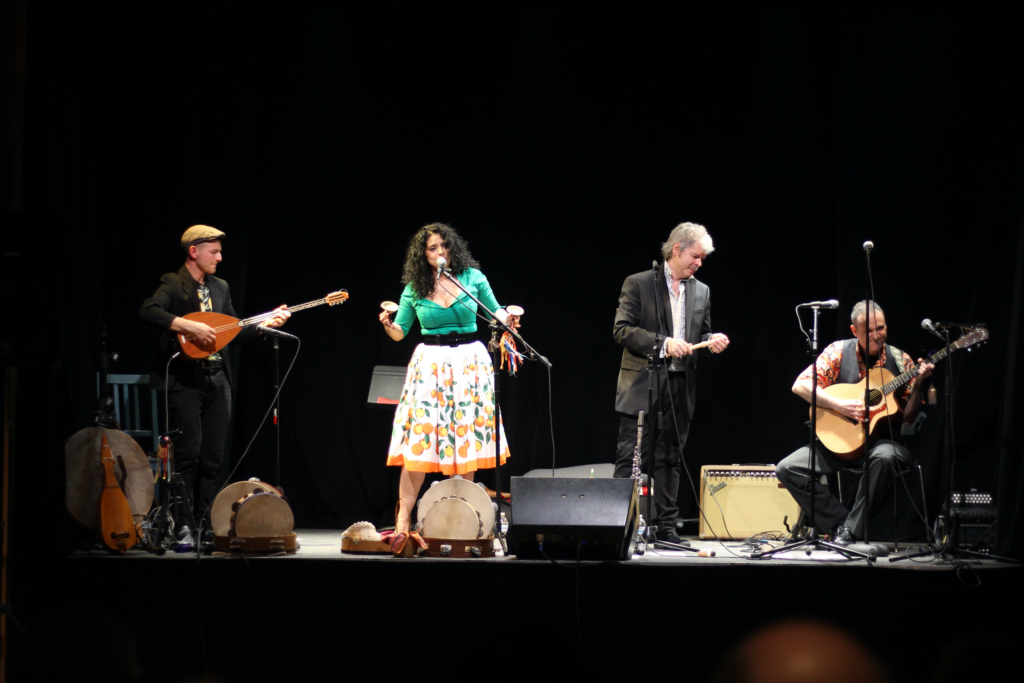 Two men playing an string instrument, one man playing a flute and a woman singing on a stage