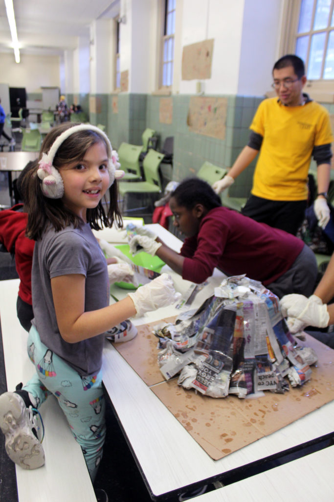 Three children sitting at a lunchroom table making a paper mache volcano with a Beacon staff member.