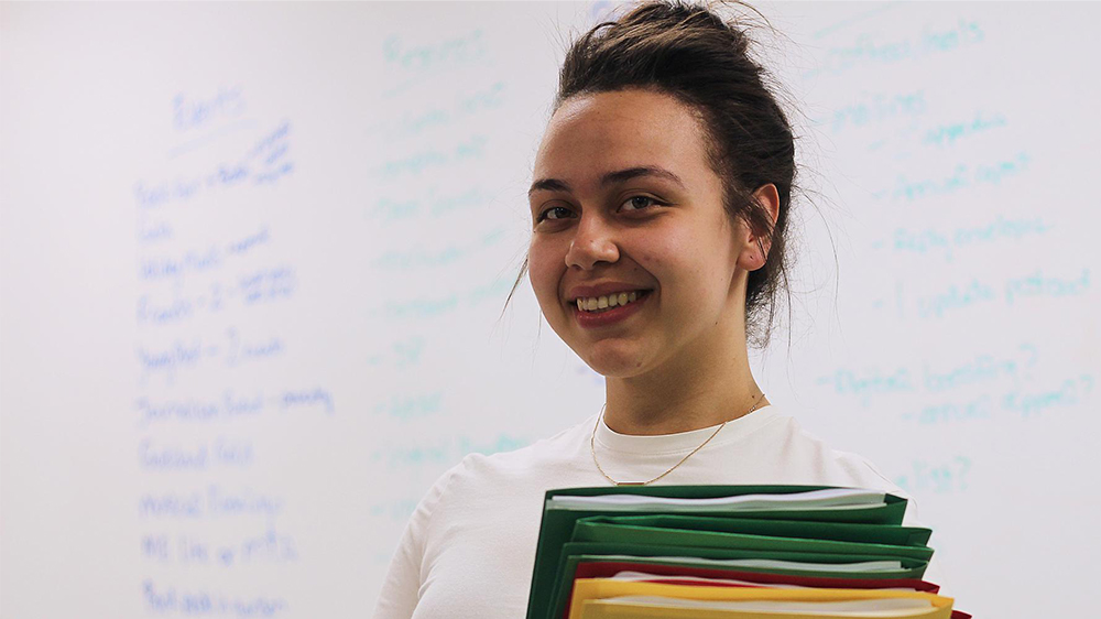 A Latina Goddard Riverside Learning to Work intern holding colorful folders stands in front of a whiteboard with writing on it.