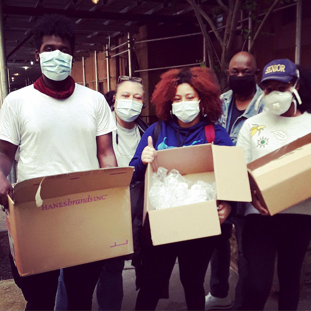 Young people wearing masks hold boxes of hand sanitizers and masks in front of a NYCHA building