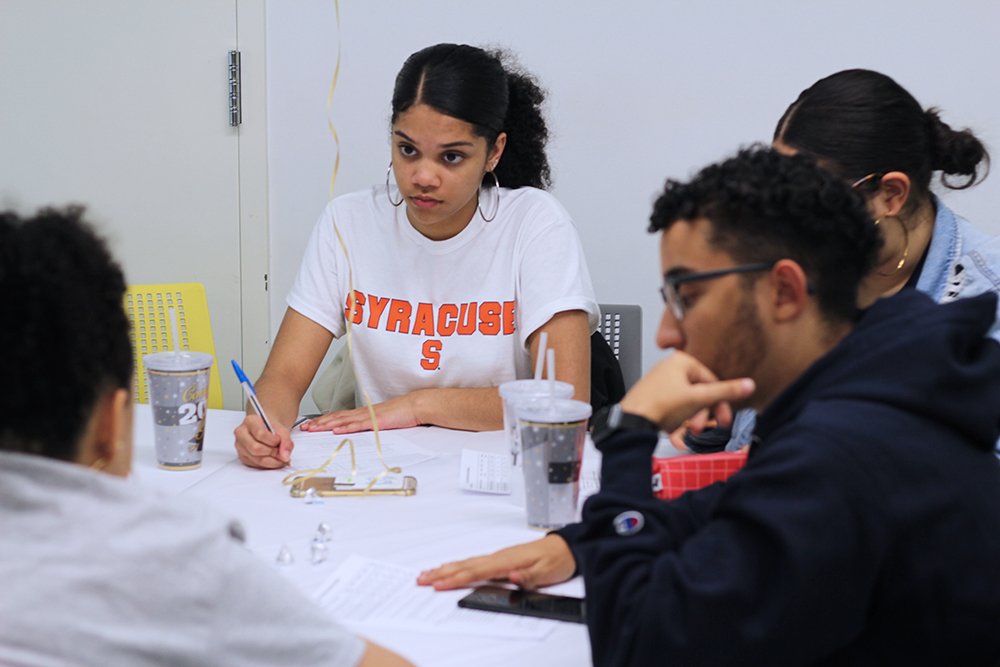 Goddard Riverside Options Center students are seated around a table doing classwork. A young woman in a Syracuse t-shirt looks across the table.