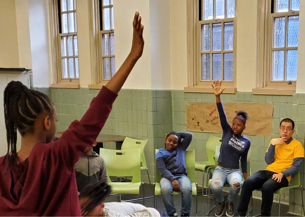 A teen standing with her back to the camera raises her hand and teens sitting facing the camera raise theirs in response.