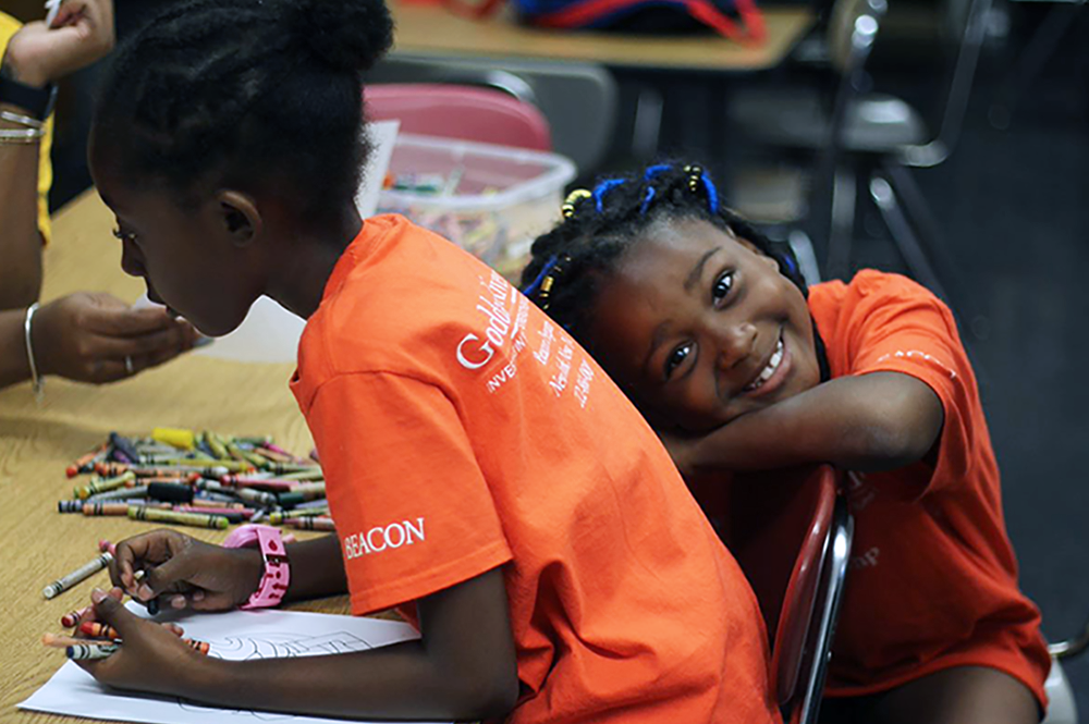 A girl in an orange Goddard Beacon summer camp shirt puts her head on her arms and smiles at the camera while her campmate makes a drawing