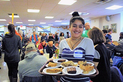 A volunteer holds a tray loaded with desserts, with tables full of people enjoying Holiday Meals in the background