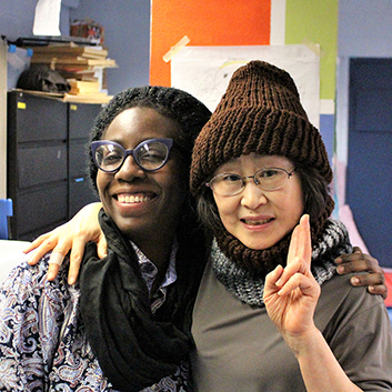 Two women, one black and one Asian, stand with an arm around each other in a Lincoln Square Senior Center knitting class