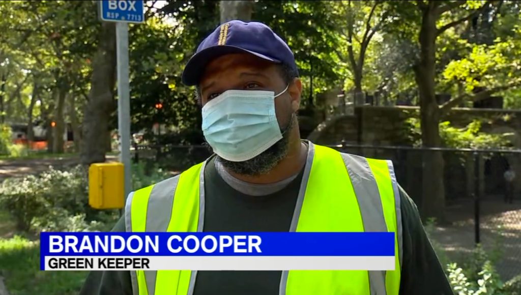 Green Keeper Brandon Cooper, wearing a face mask and a bright green vest, is interviewed by a TV reporter