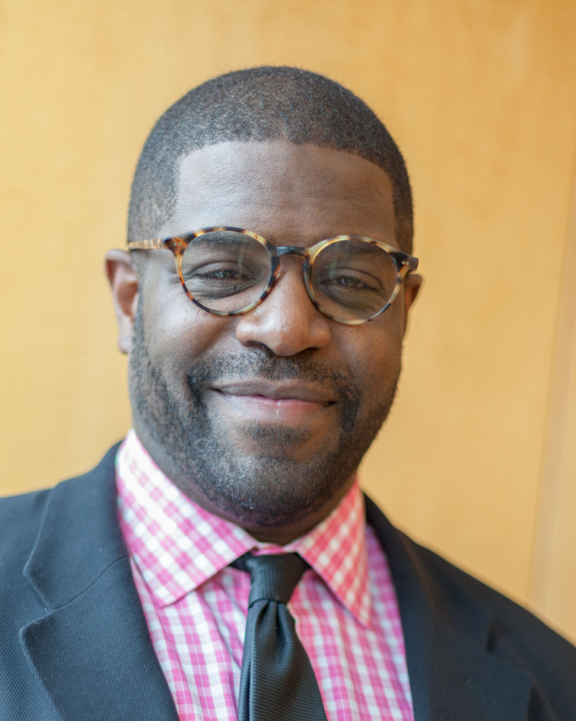 Headshot of Goddard Riverside Executive Director Roderick L. Jones wearing a pink and white checked shirt and gray suit and tie