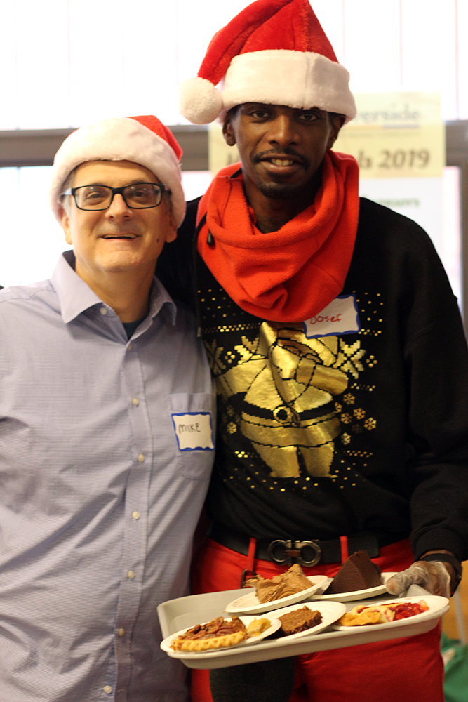 Two men in Santa hats stand together, one holding a tray of pastries