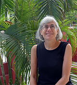A woman with chin-length silver hair and glasses sits in front of a lush palm tree