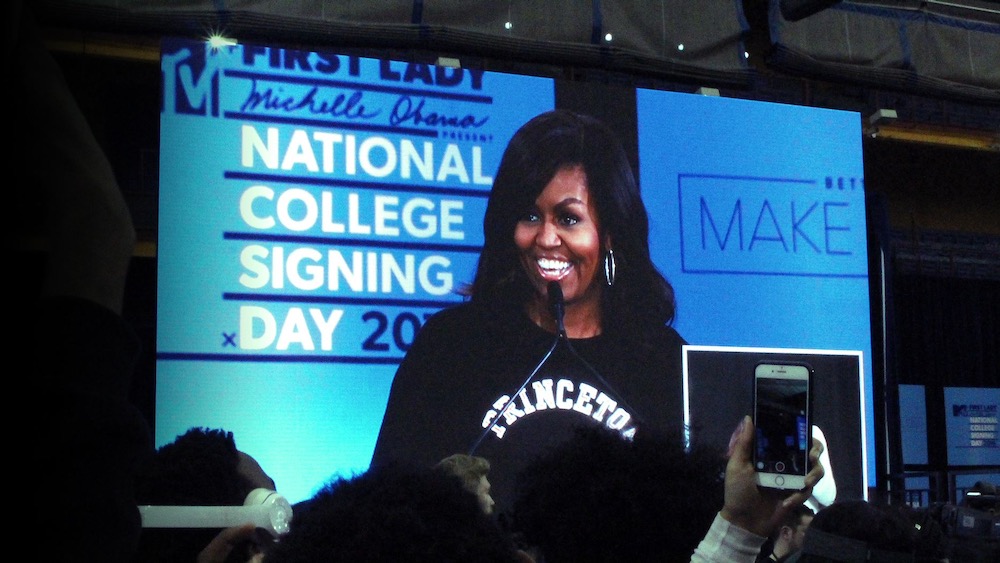 Michelle Obama speaks on a giant video screen in an auditorium, wearing a black Princeton sweatshirt with white lettering, while young people raise their cellphones to take photos.