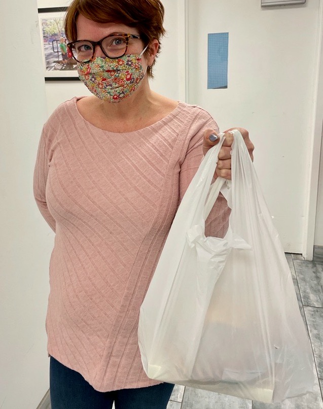 A woman in a pink sweater, wearing a mask, holds a delivery meal in a white plastic bag