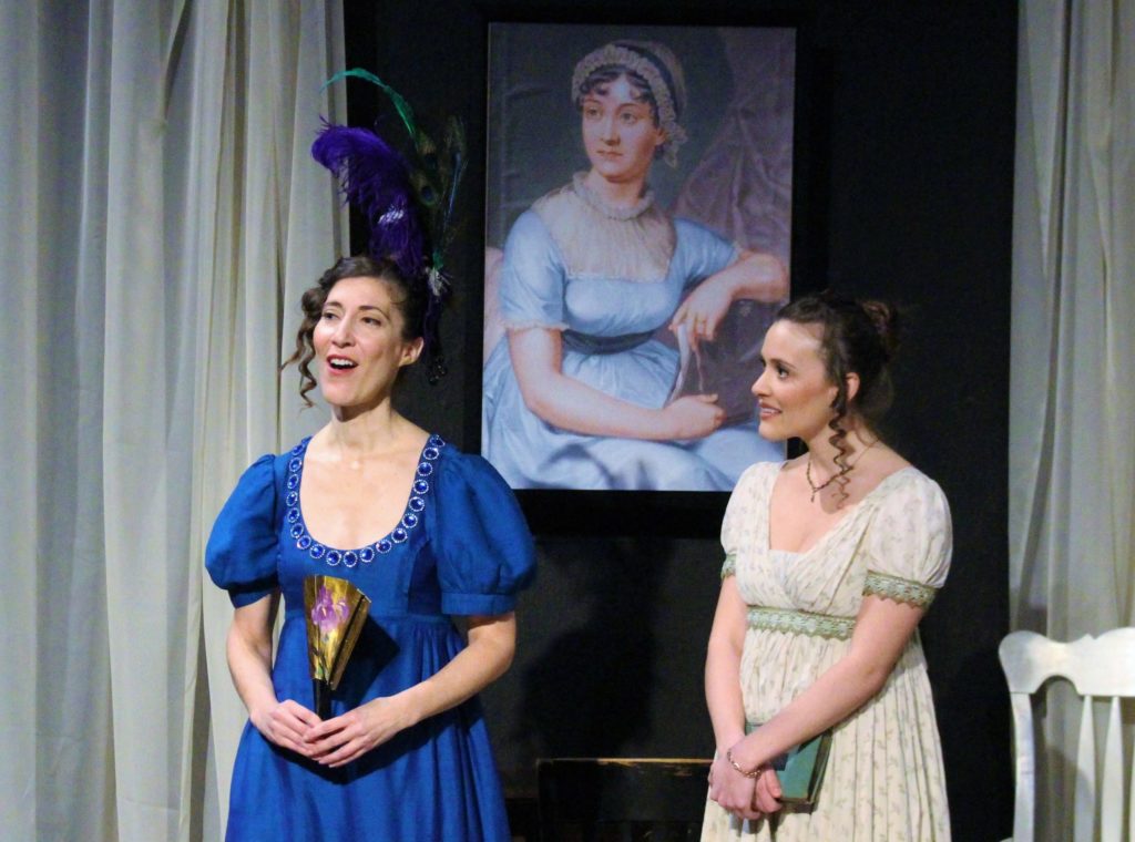 two women standing in Regency period clothing in front of a Jane Austen portrait
