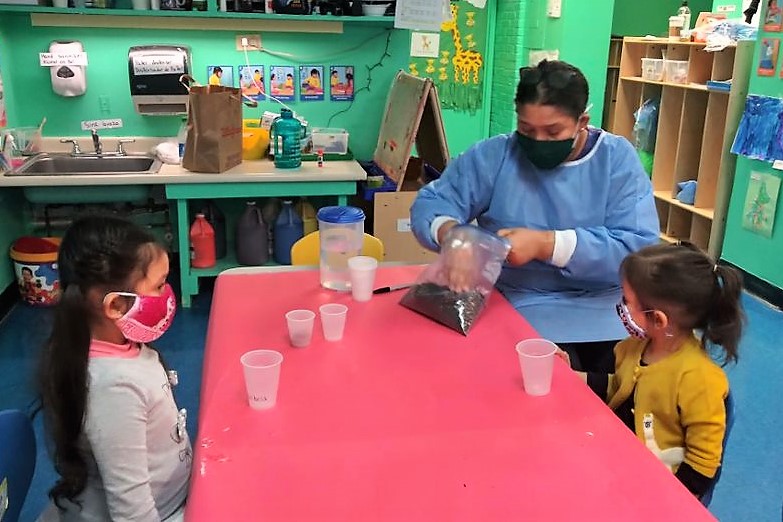 Two children sit at a table with plastic cups in front of them while an adult digs into a bag of soil