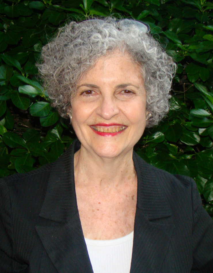 Susan Richman, with curly silver hair and bright red lipstick, standing in front of a background of dark leaves