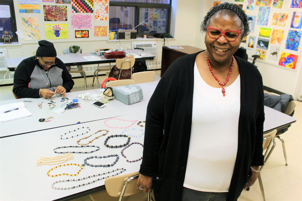 Two women in an art classroom creating jewelry. One of the woman is standing with a handmade necklace and smiling.
