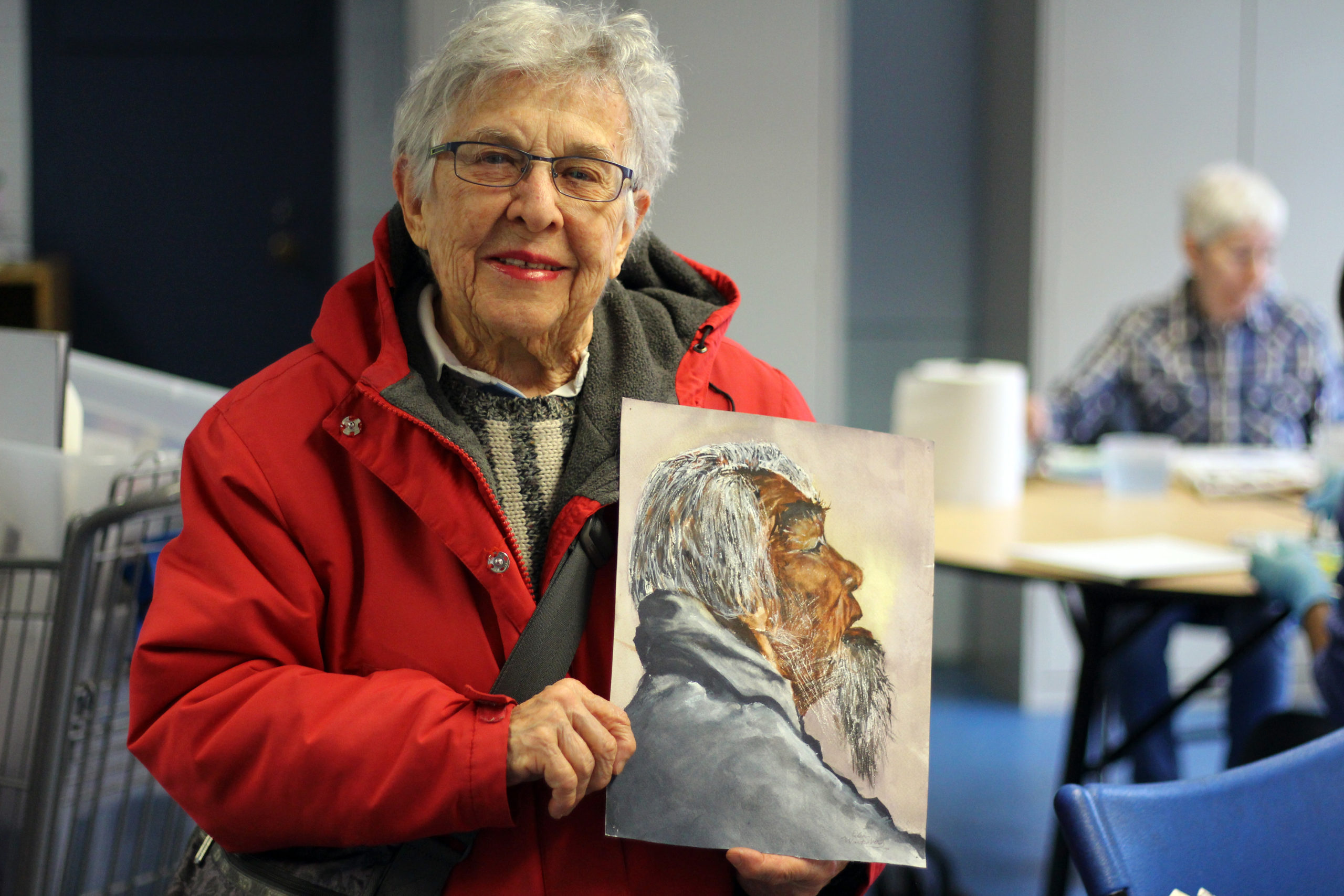 An older woman in a red jacket holds up a painting she's done