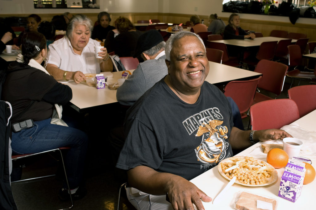 A man sitting in a dinning room with other seniors, smiling with food in front of him. 
