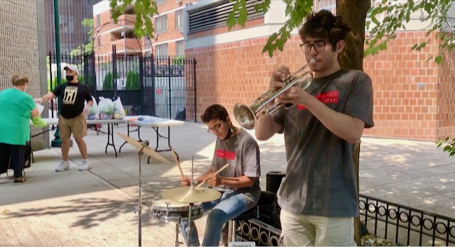 Two teen boys play trumpet and violin on a city sidewalk with a table filled with bags of food in the background
