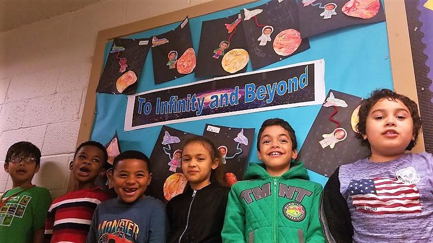 Six kids standing in front of a bulletin board.