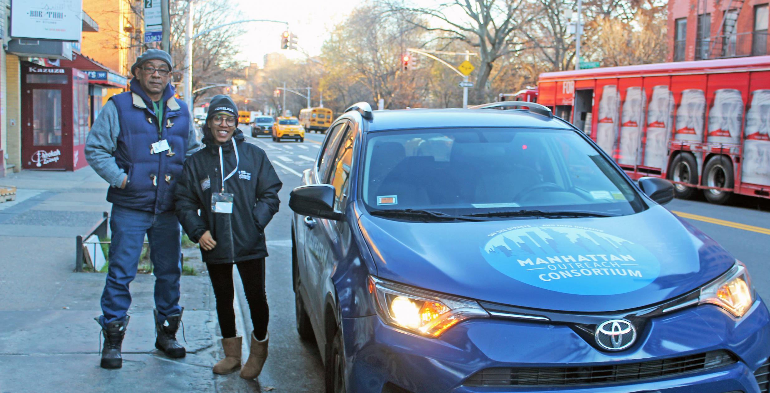 Two people dressed for cold weather stand next to a small SUV labeled Manhattan Outreach Consortium as dawn breaks over the city