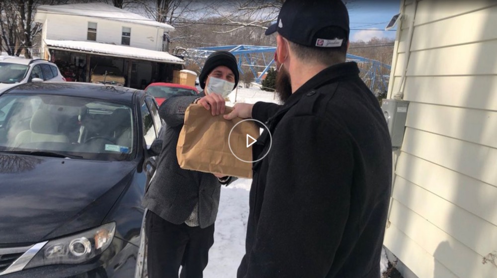 Screenshot of a man in a snowy landscape standing next to a car and handing another man a paper bag presumably containing food