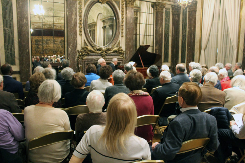 A crowd of people sitting listening to a classical music artist on a piano. 