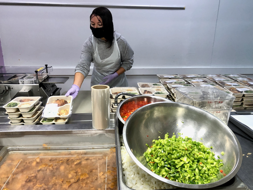 Photo of a metal bowl of chopped green peppers. In the background, a woman wearing a hairnet and masks stacks meals ready for home delivery .