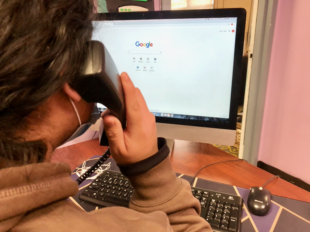 A young man's head is seen from behind as he holds a phone to his ear while looking at a computer screen