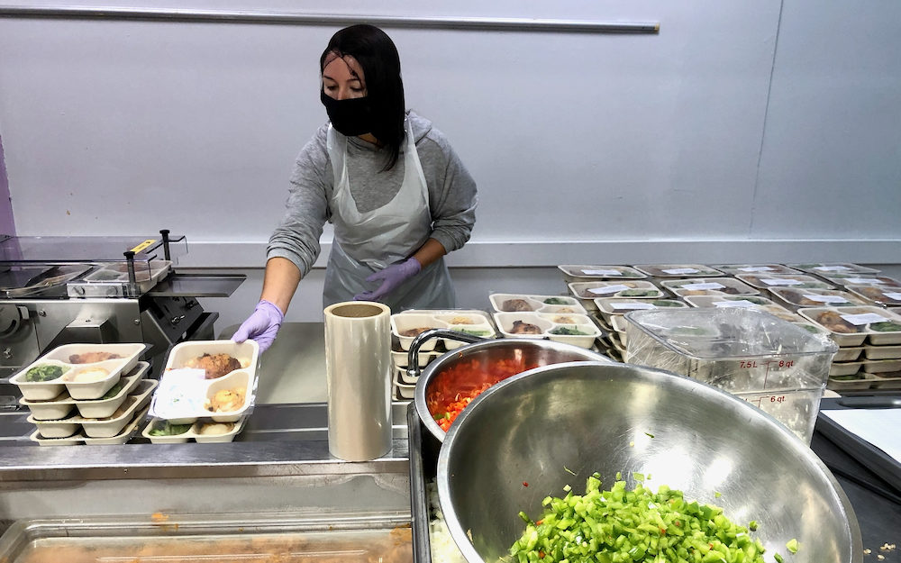 Photo of a metal bowl of chopped green peppers. In the background, a woman wearing a hairnet and masks stacks meals ready for home delivery .