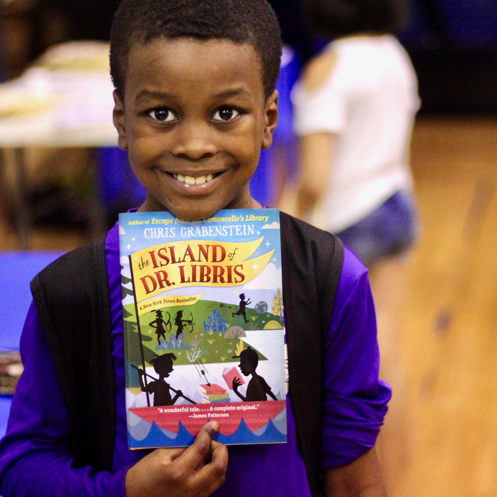 A smiling boy holds a book in the dining room and gymnasium at Lincoln Square Neighborhood Center
