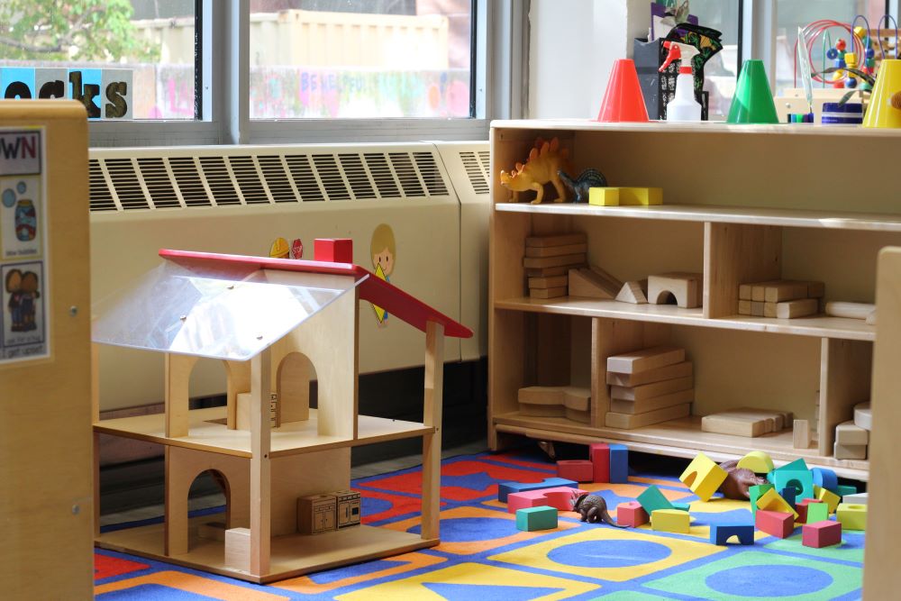 A play area featuring a soft floor mat, a simple wooden house and colorful blocks