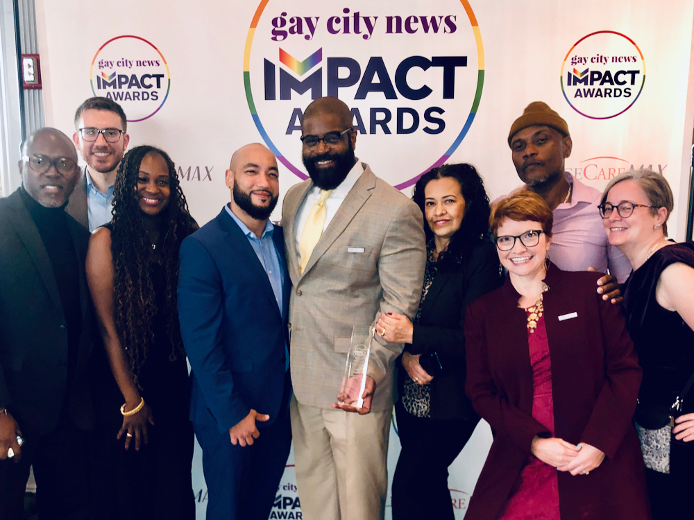 Several people pose for a photo in front of a Gay City News Impact Awards backdrop