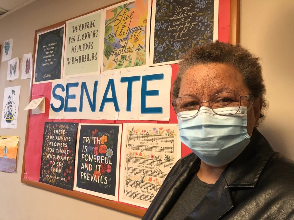 A woman stands next to a bulletin board with the word SENATE in large letters. Also featured is the quote "Work is Love made Visible."