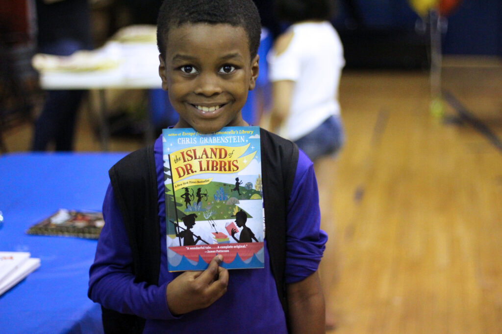 A smiling boy holds a book in the dining room and gymnasium at Lincoln Square Neighborhood Center.