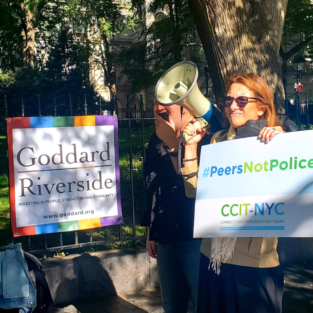 A woman holding a blow horn, while holding a sign at a rally with a Goddard Riverside banner behind her.