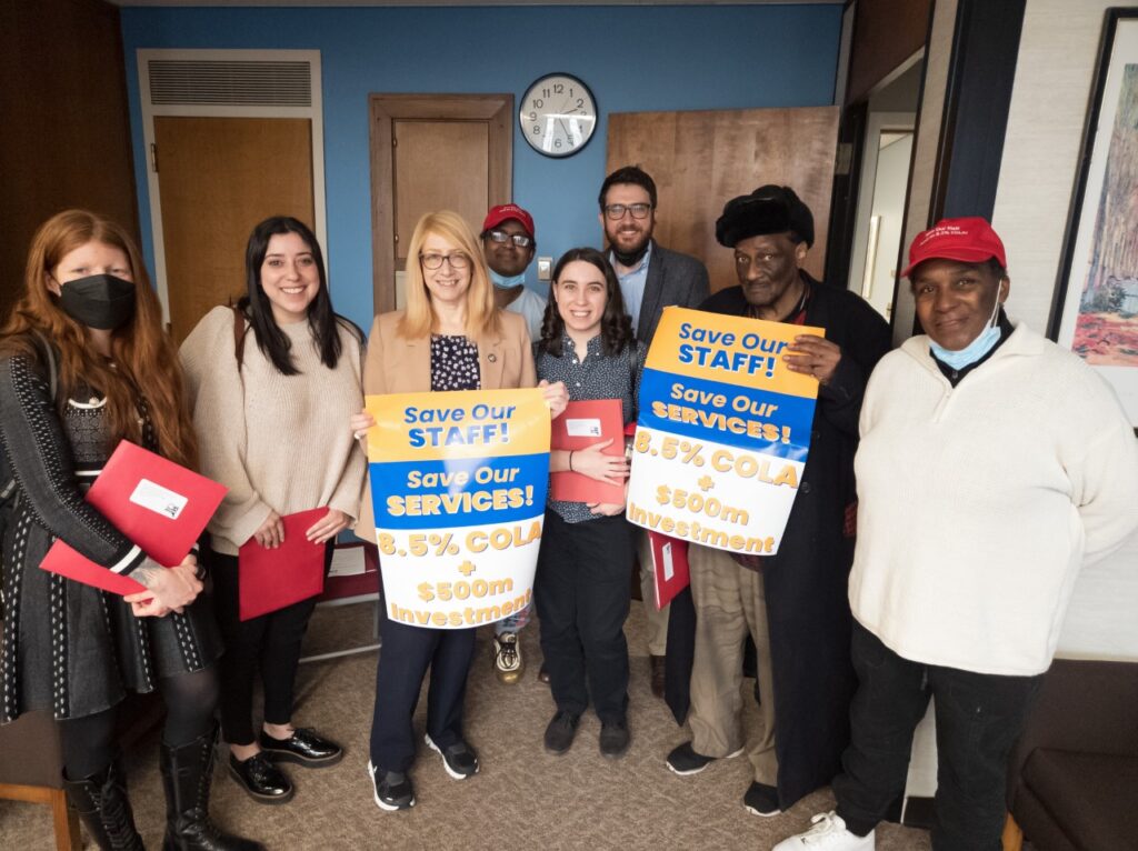 Several people pose with NY Assembly Member Linda B. Rosenthal. Two hold signs saying "Love our staff! Save our services! 8.5% COLA and $500 million investment."