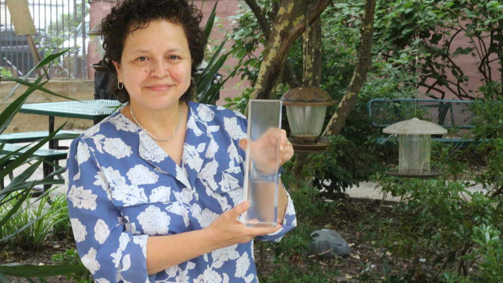 A woman wearing a blue-gray shirt with large white flowers stands in a garden holding a lucite award statuette