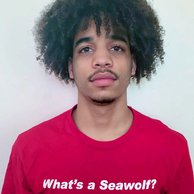 A young man wearing a red SUNY Stonybrook shirt with the slogan "What's a Seawolf?"