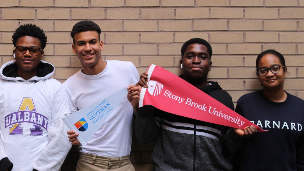Four young people stand outside against a tan-colored brick wall wearing sweatshirts or carrying pennants of U Albany, Guttman, Stony Brook and Barnard