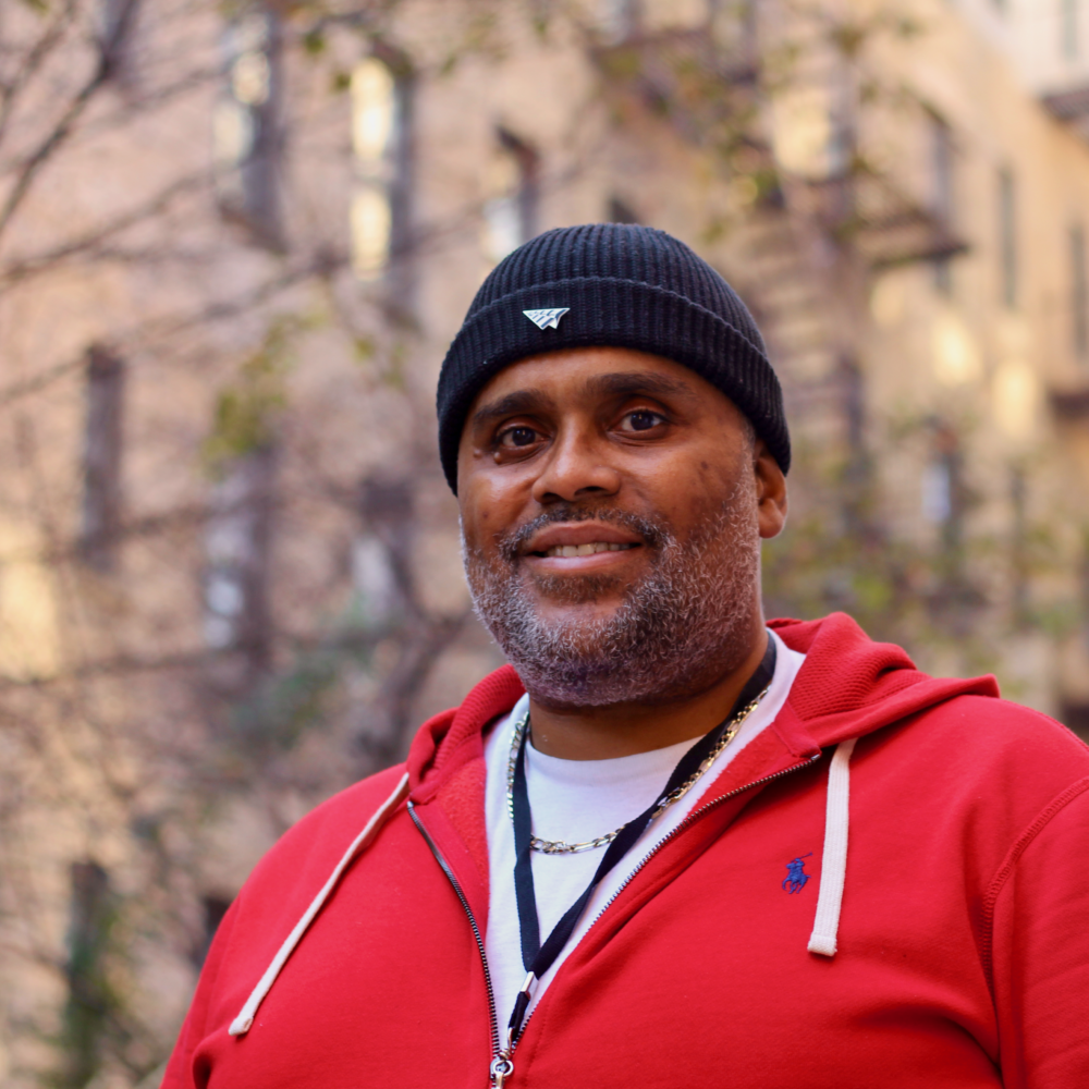 A man in a red zipfront hoodie and dark knit hat poses for a photo with an out-of-focus apartment building in the background