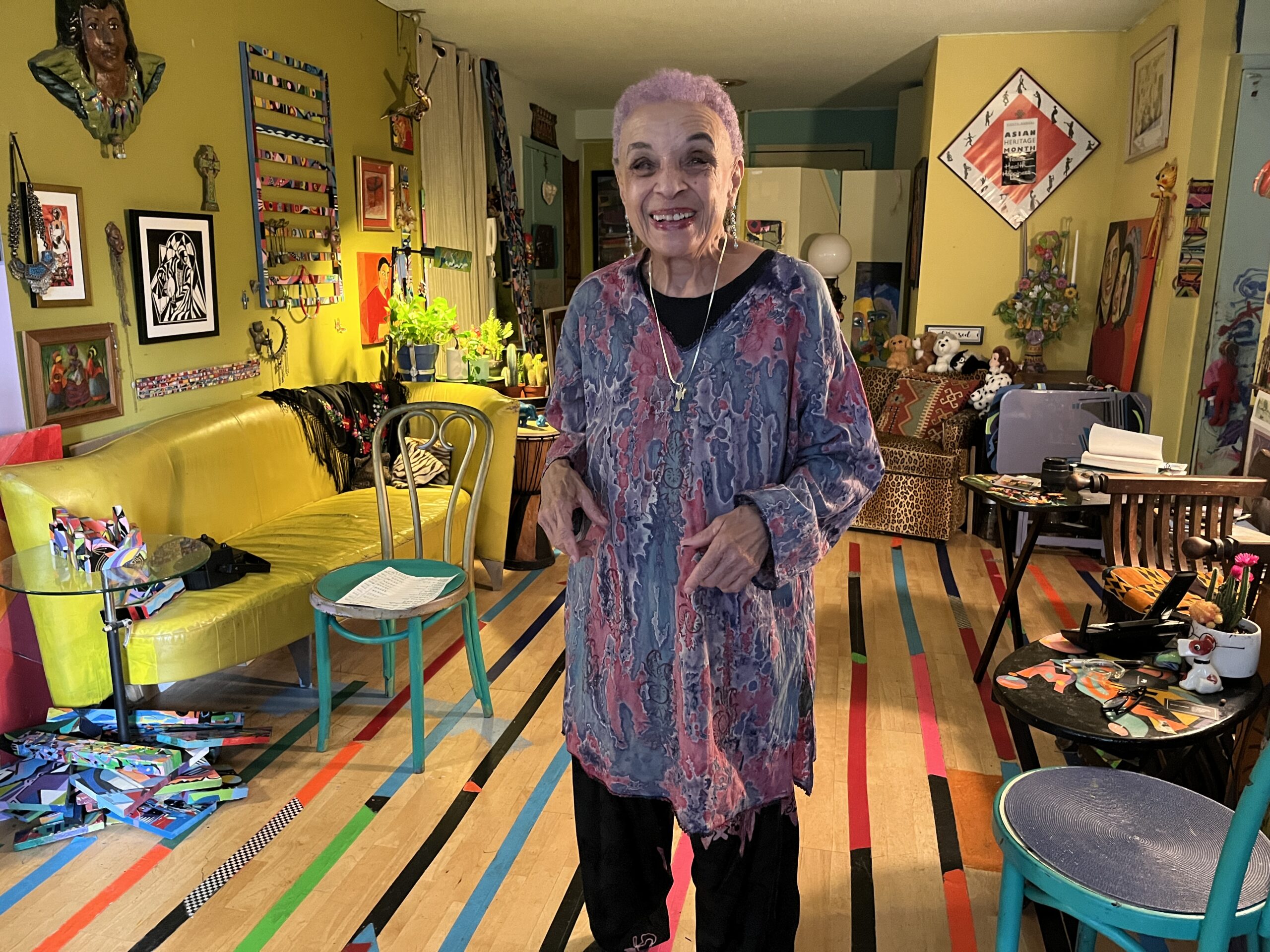 Nancy Brandon stands smiling in a brightly colored and eclectically decorated room, with a yellow sofa, various chairs, wall art, and vibrant patterns on the floor and walls.
